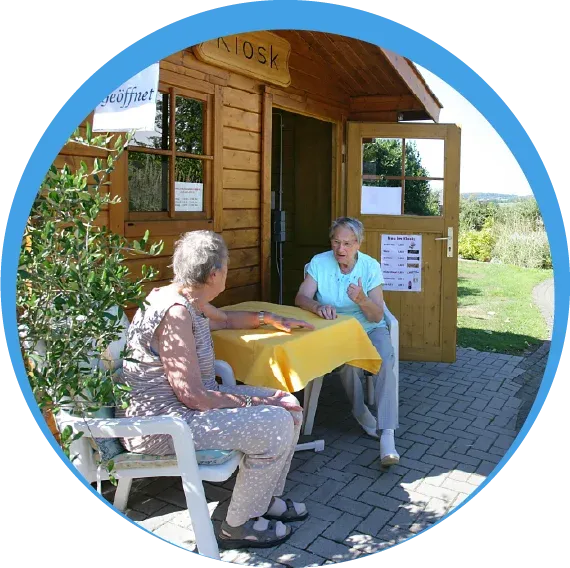 Zwei Seniorinnen sitzen an einem gelb gedeckten Tisch vor einem rustikalen Holzhaus im Außenbereich der Wohnanlage Appenhagen.